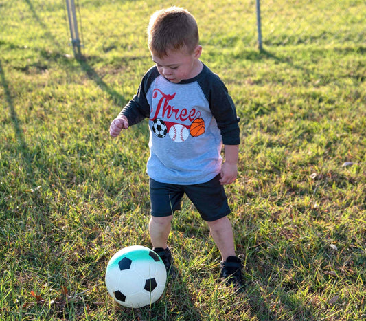 May include: A young child wearing a gray and black baseball shirt with the word &#39;Three&#39; and images of a soccer ball, baseball, and basketball. The child is standing on a grassy field and kicking a soccer ball.